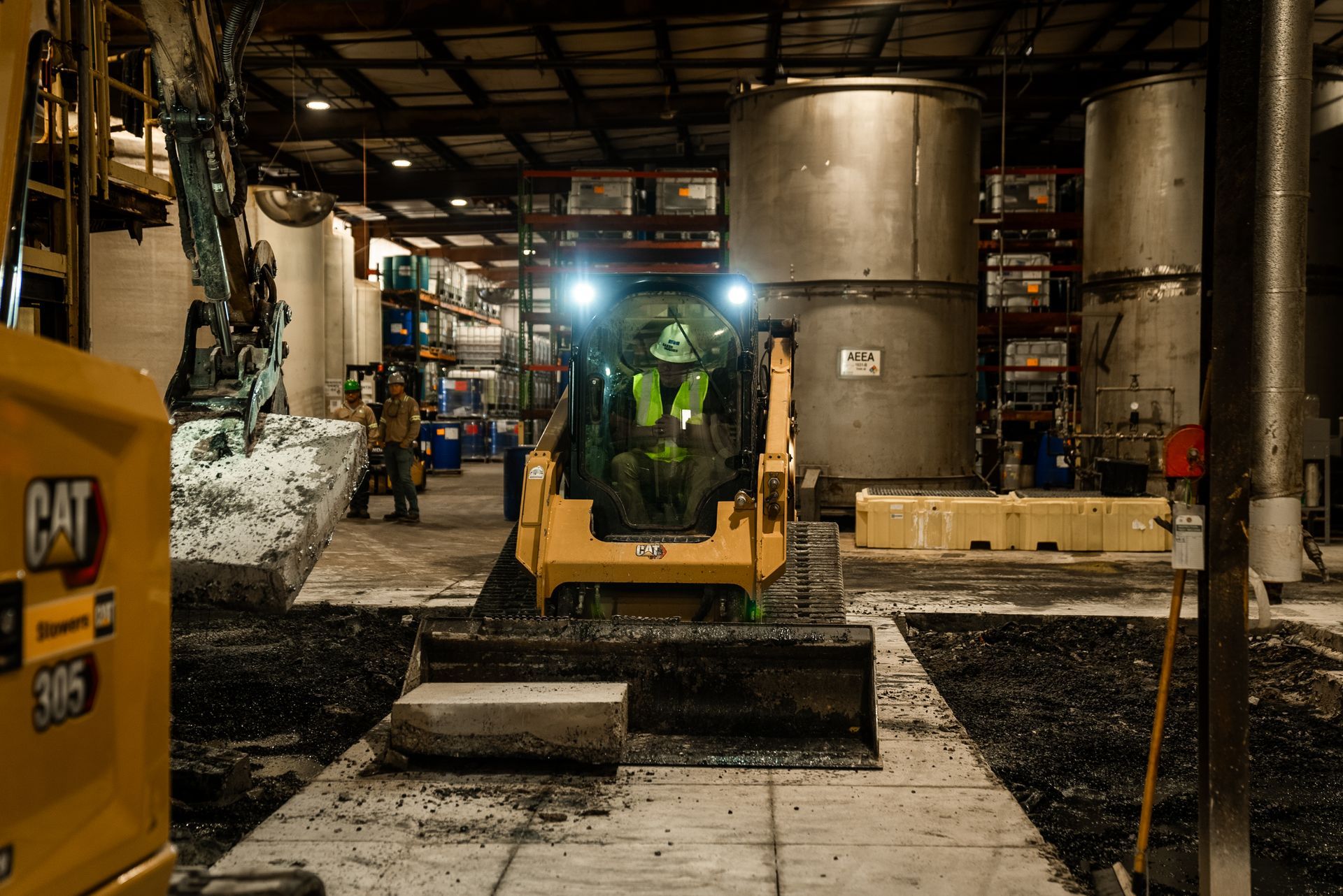 Skid steer loader in warehouse, operator inside, working near large metal tanks and equipment.