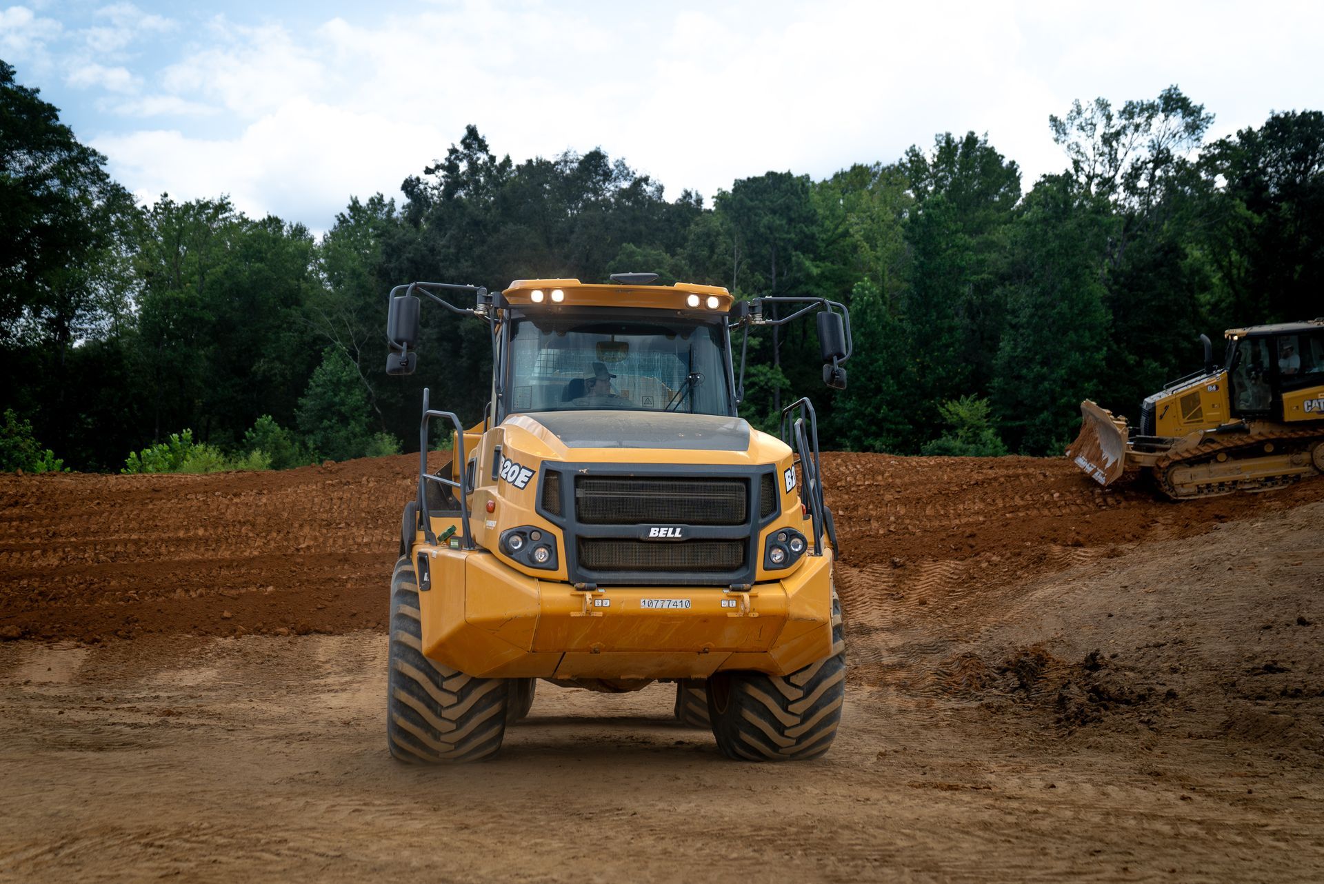 Yellow construction machine on dirt, with trees in the background. Another machine to the right.
