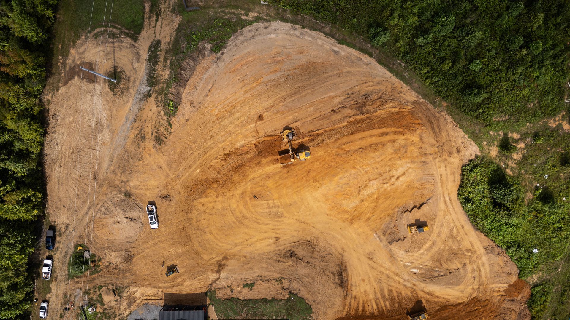 Aerial view of a large pile of wood chips with machinery, near a forest edge and parked vehicles.