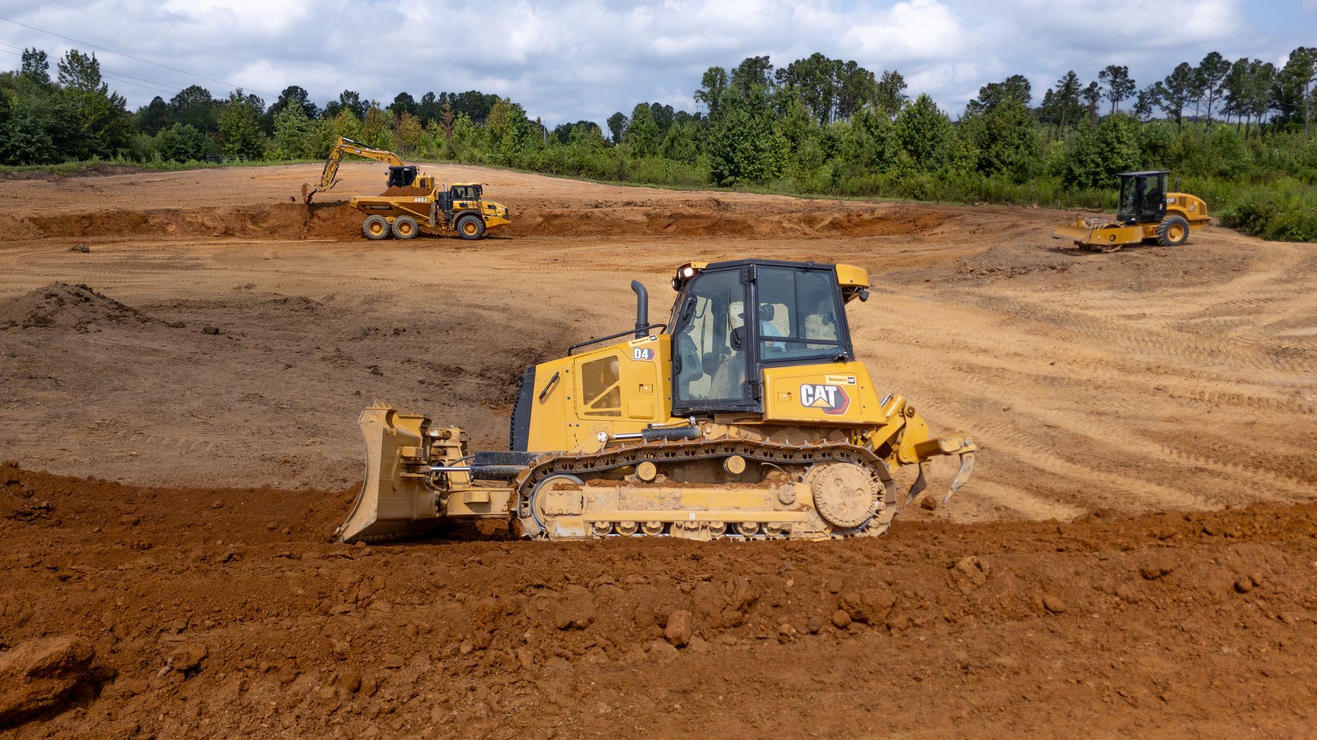 Yellow bulldozer pushes earth at a construction site; other machines work in the background under a blue sky.