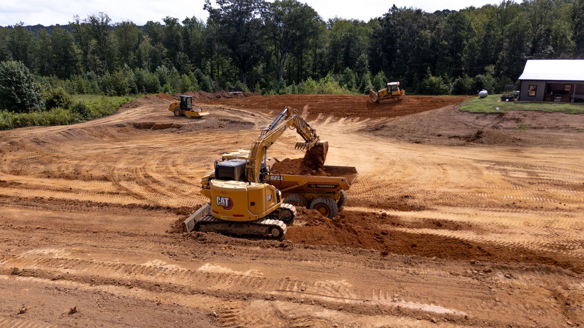 An excavator loading a dump truck at a construction site. Dirt is piled up. Green trees in the background.