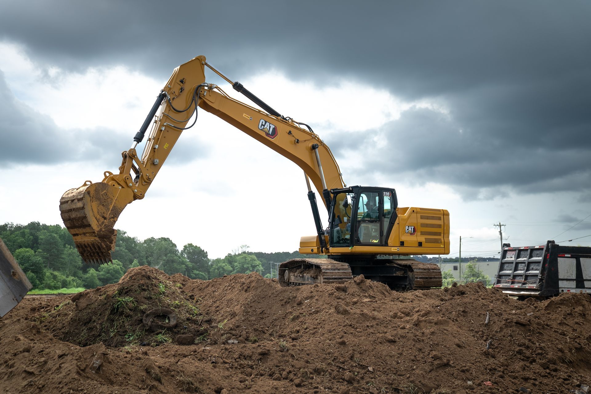 Yellow Caterpillar excavator scoops earth from a dirt pile under a cloudy sky.