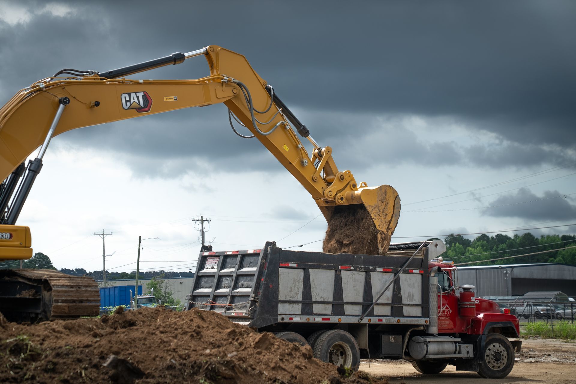 Yellow excavator loading dirt into a red dump truck, under a cloudy sky.