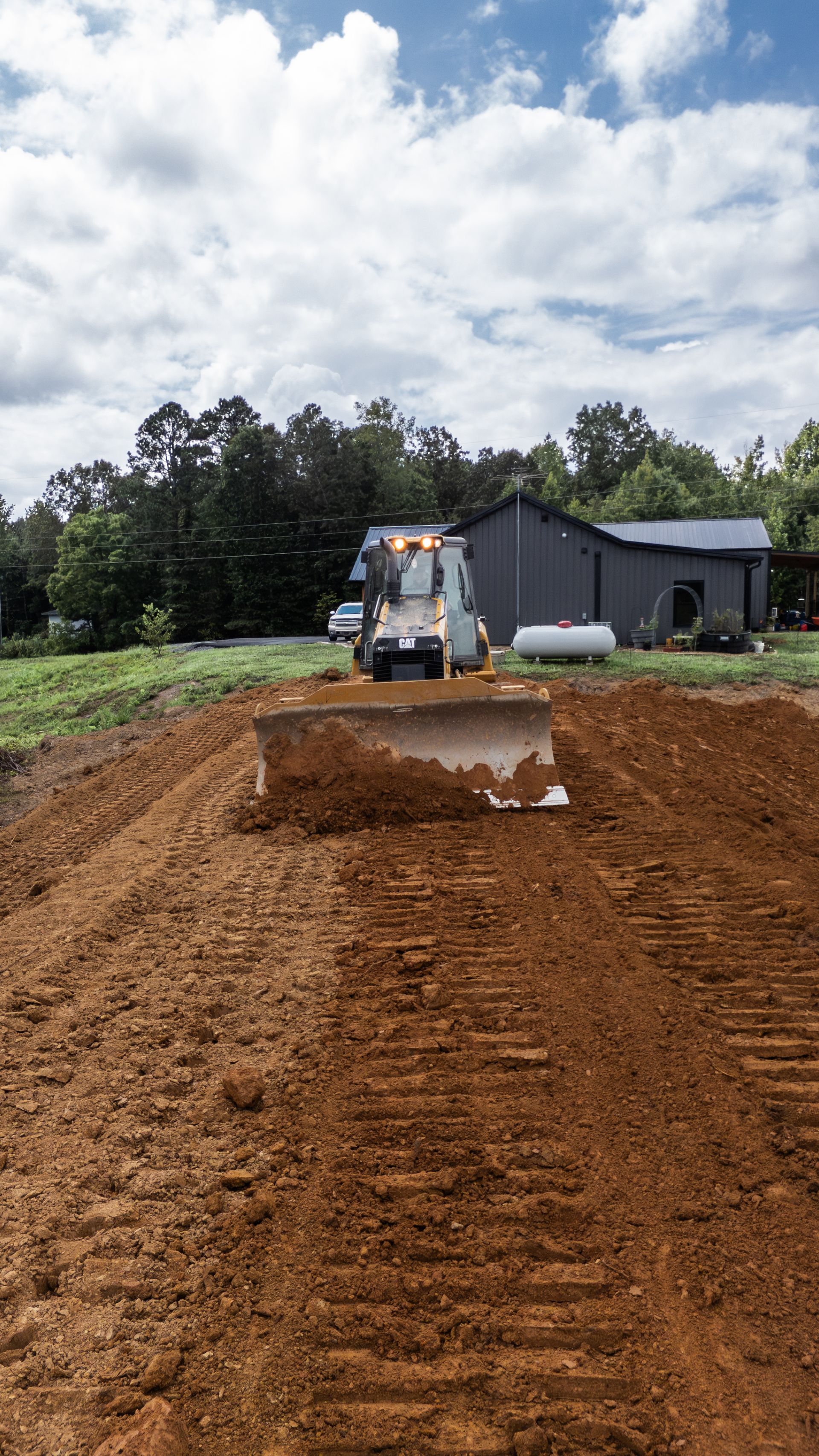 A bulldozer pushes soil, forming a berm in a field near a barn under a cloudy sky.