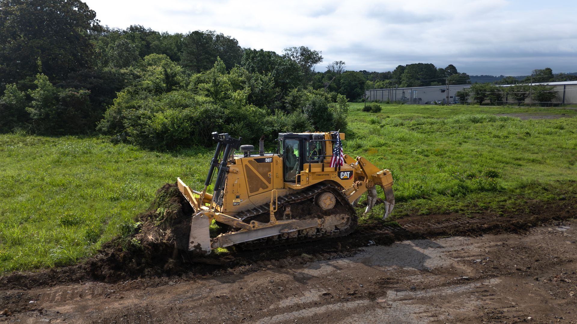 Yellow bulldozer clearing earth in a grassy field near trees and a building.
