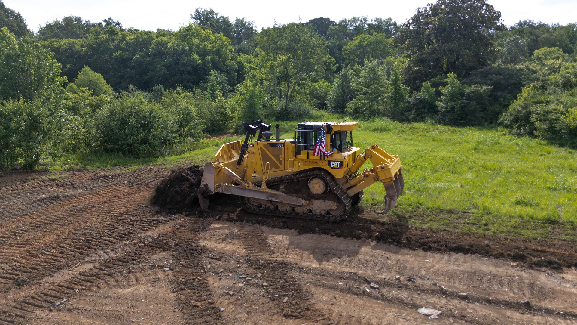 Yellow bulldozer pushing earth on a grassy area, with trees in the background on a sunny day.
