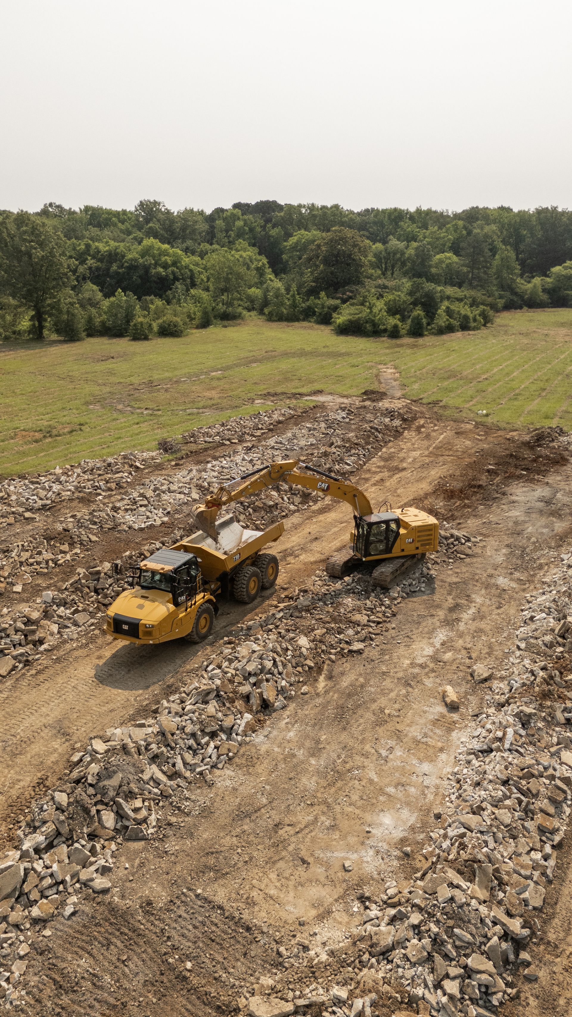Two yellow construction vehicles on a rocky, dirt ground; trees and sky in background.