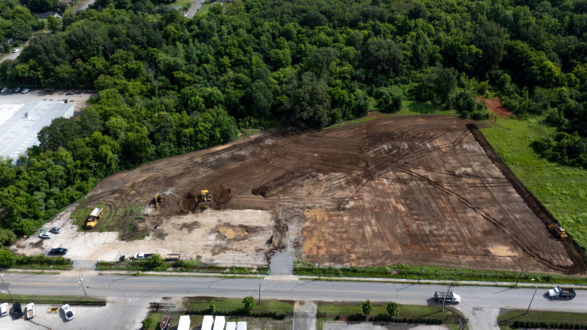 Construction site, earthmovers working on clearing a hillside; road in foreground, trees in background.
