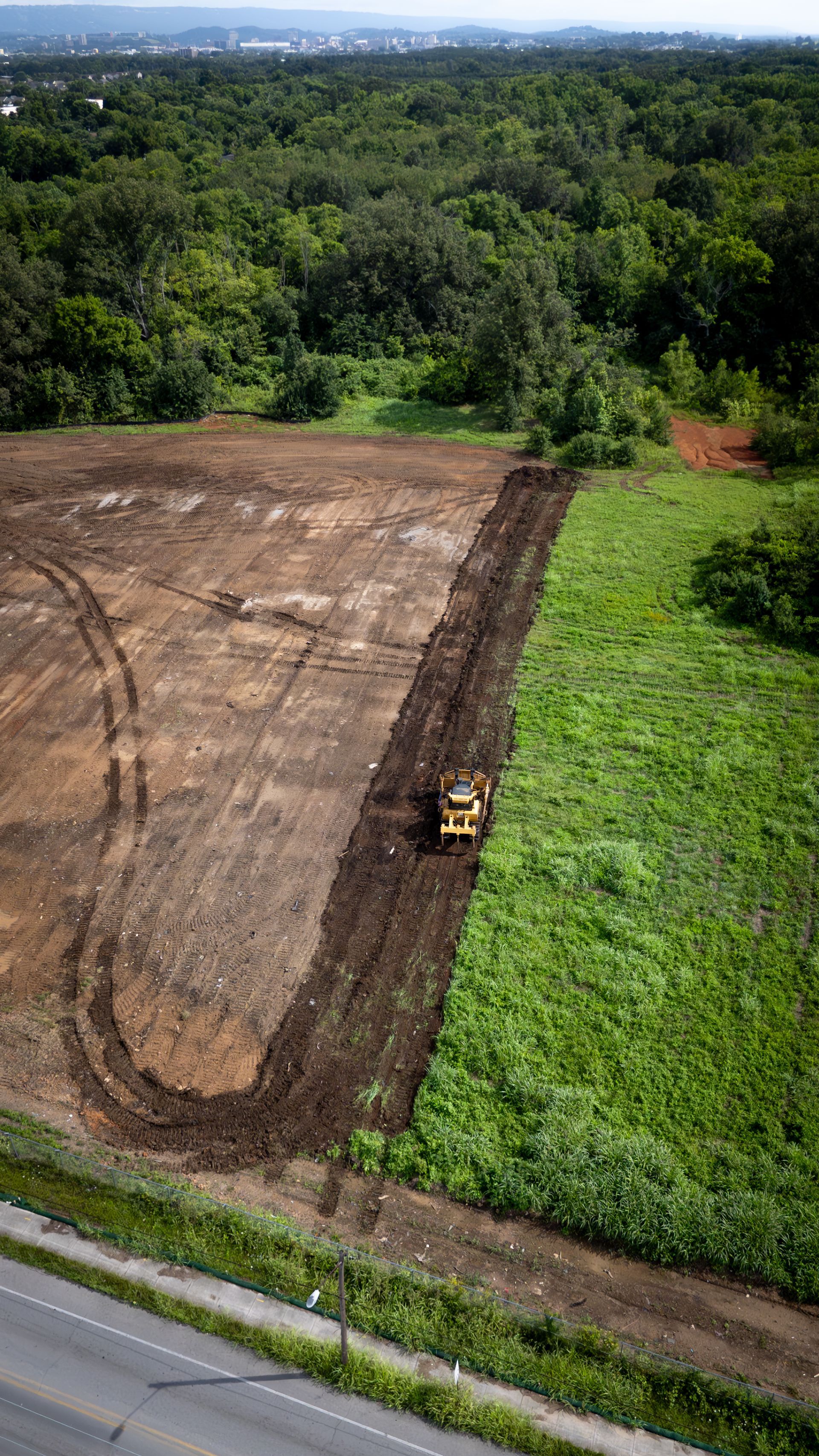 Aerial view: Bulldozer clearing land alongside a green field and road, with a treeline in the background.