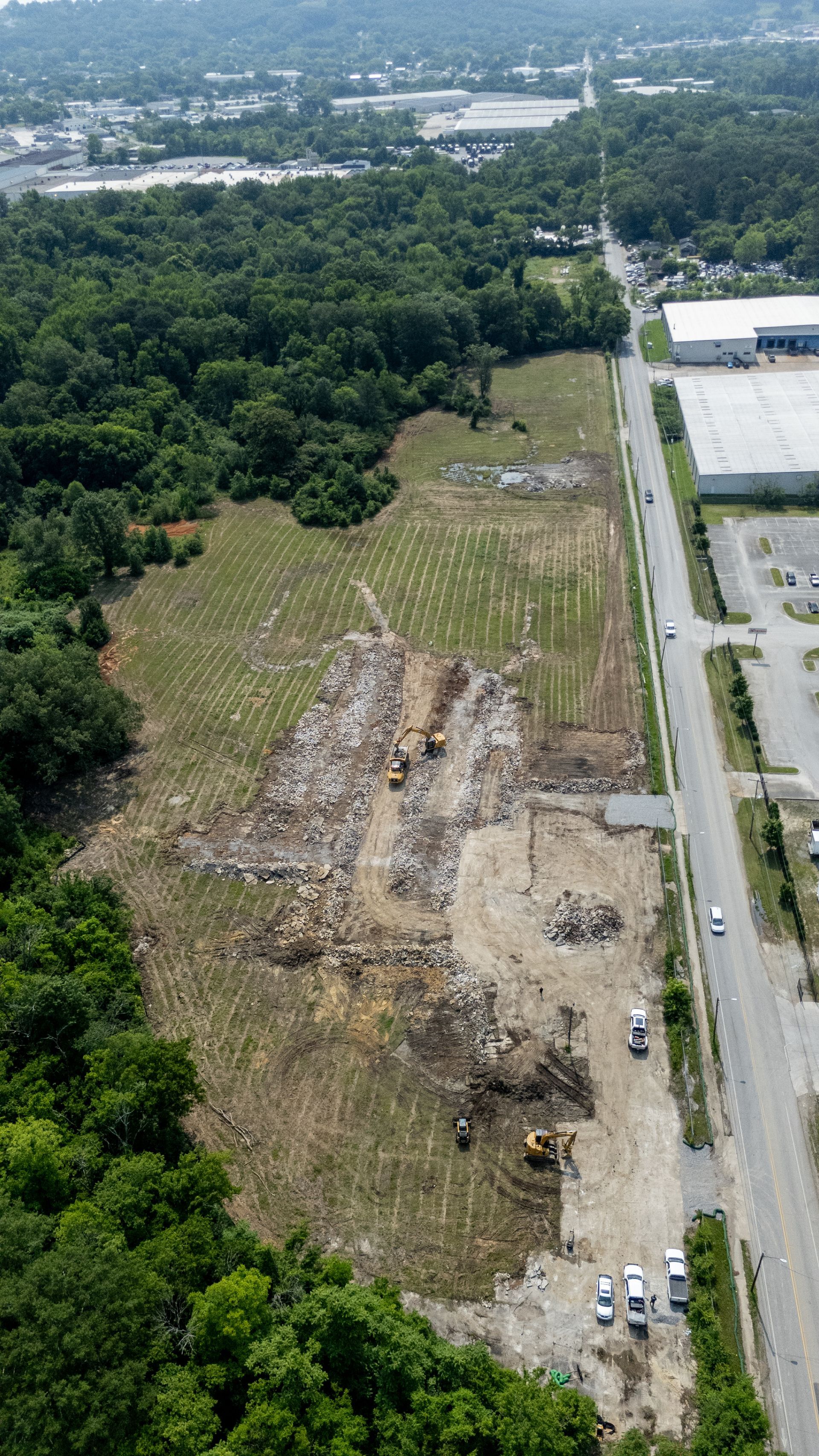 Aerial view of a cleared construction site with heavy machinery, adjacent to a road and forest.