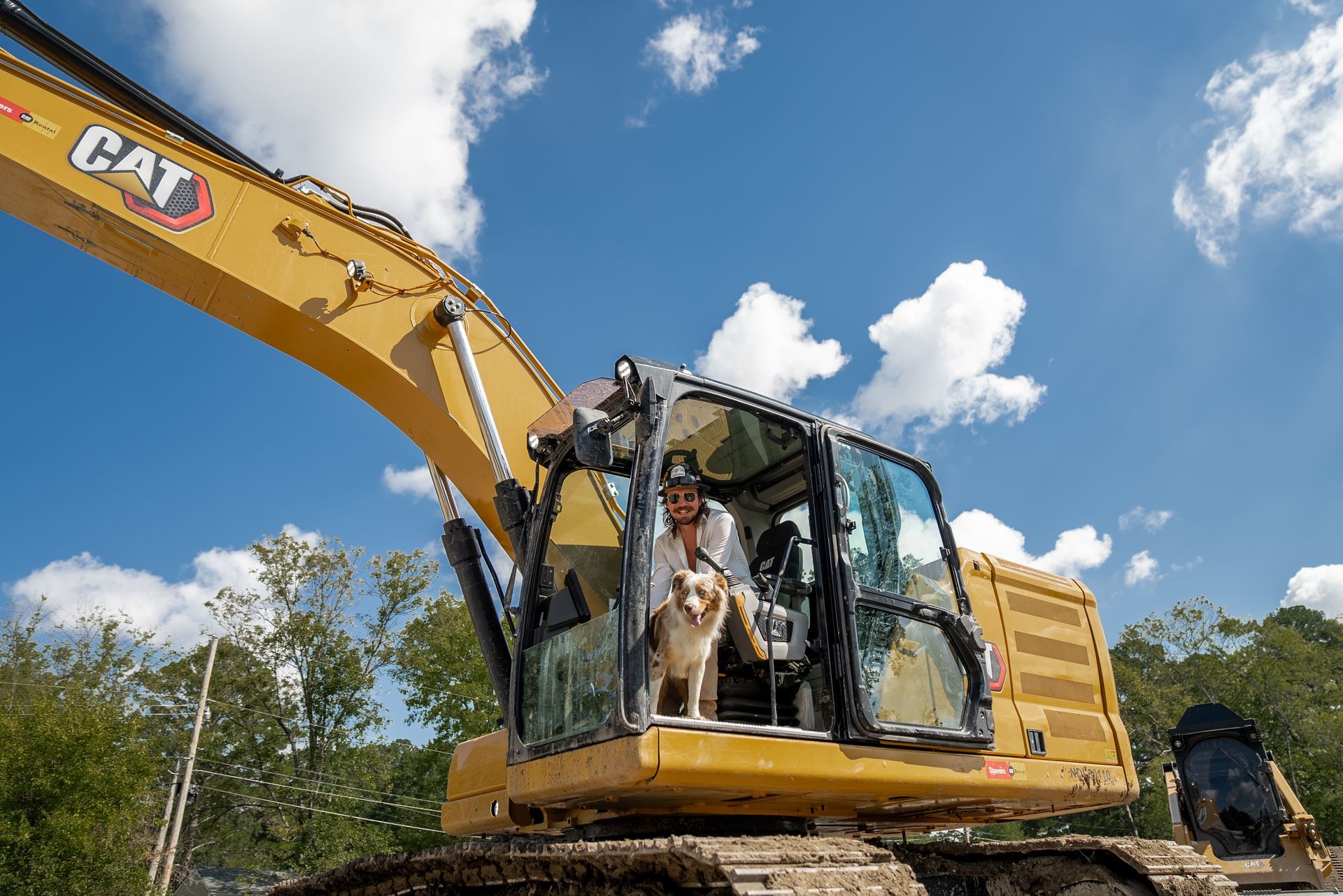 A person and dog in a yellow CAT excavator cab under a blue sky.
