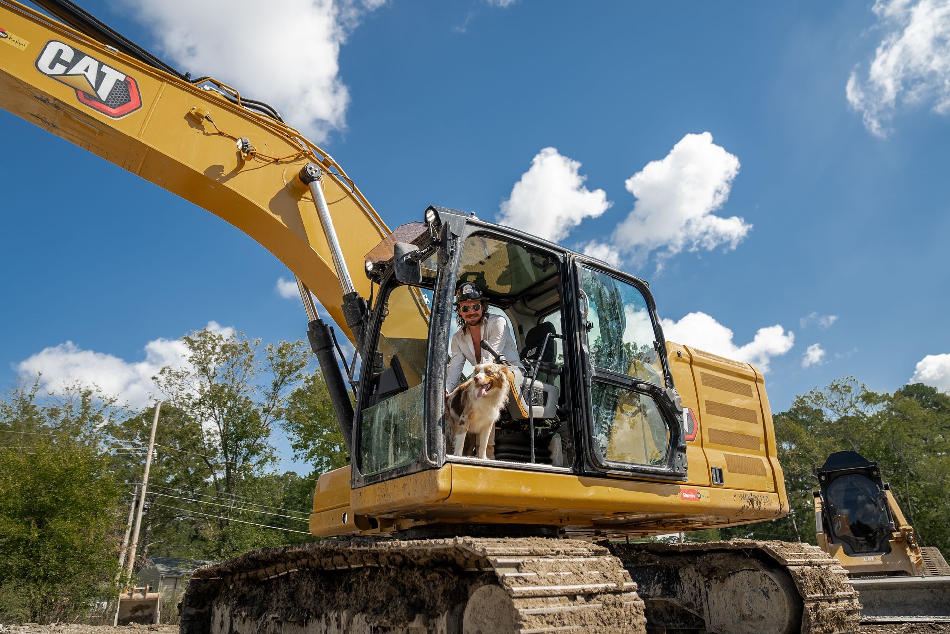 A person and dog sit in the cab of a yellow CAT excavator against a blue sky.