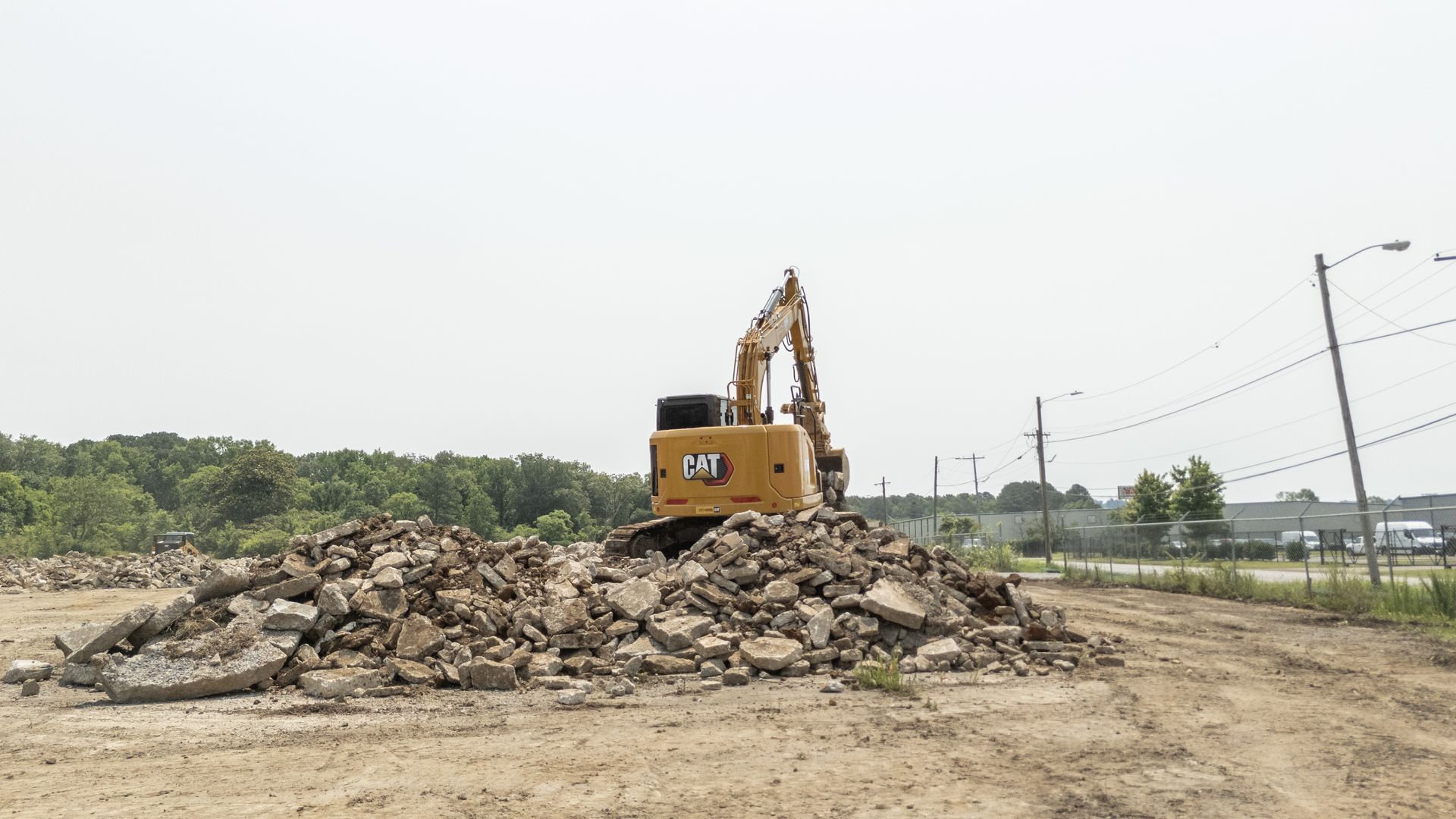 Yellow excavator on a pile of rubble on a dirt lot, trees and power lines in the distance.