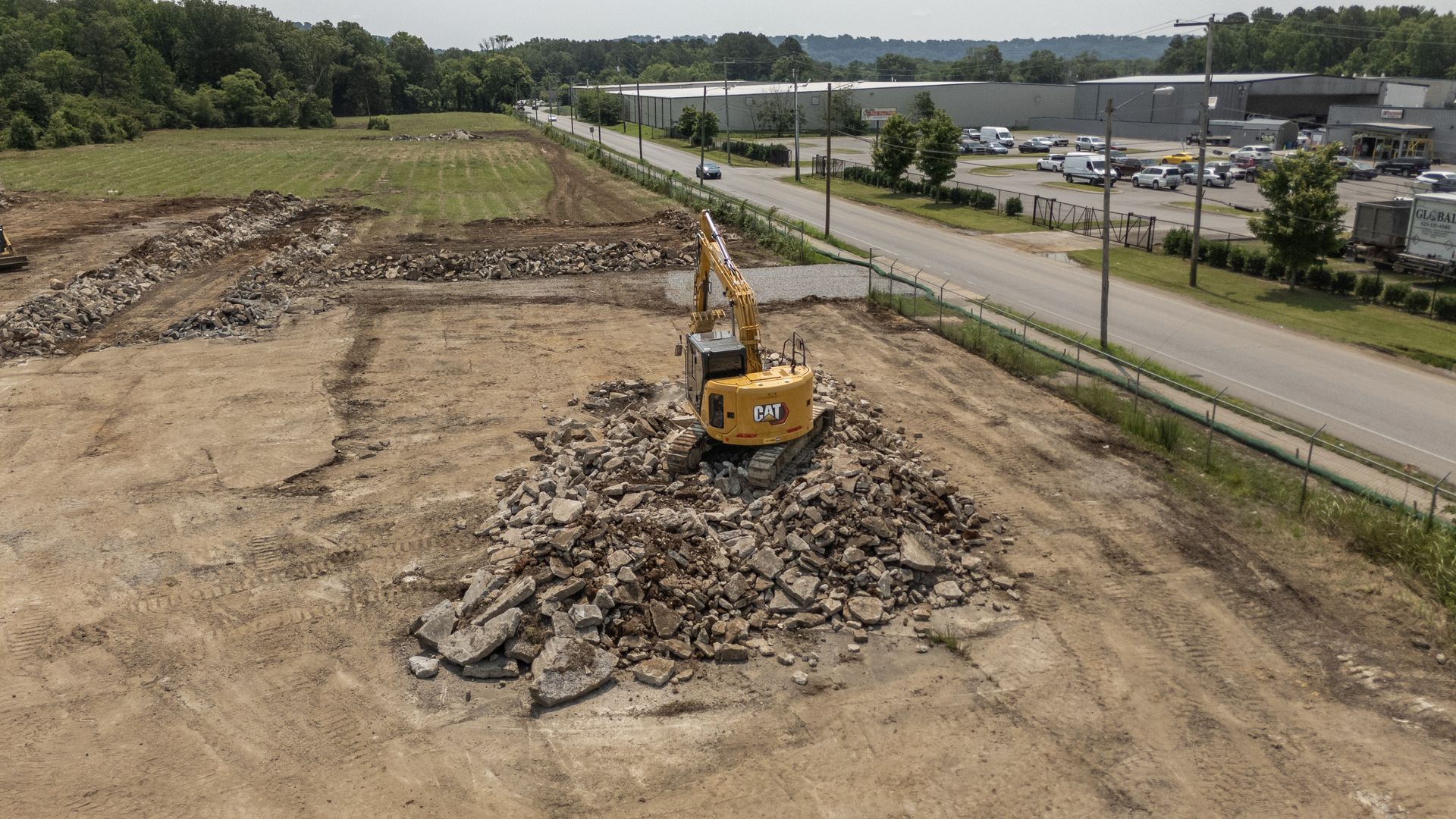 Excavator demolishing debris on a construction site near a road and parked vehicles.
