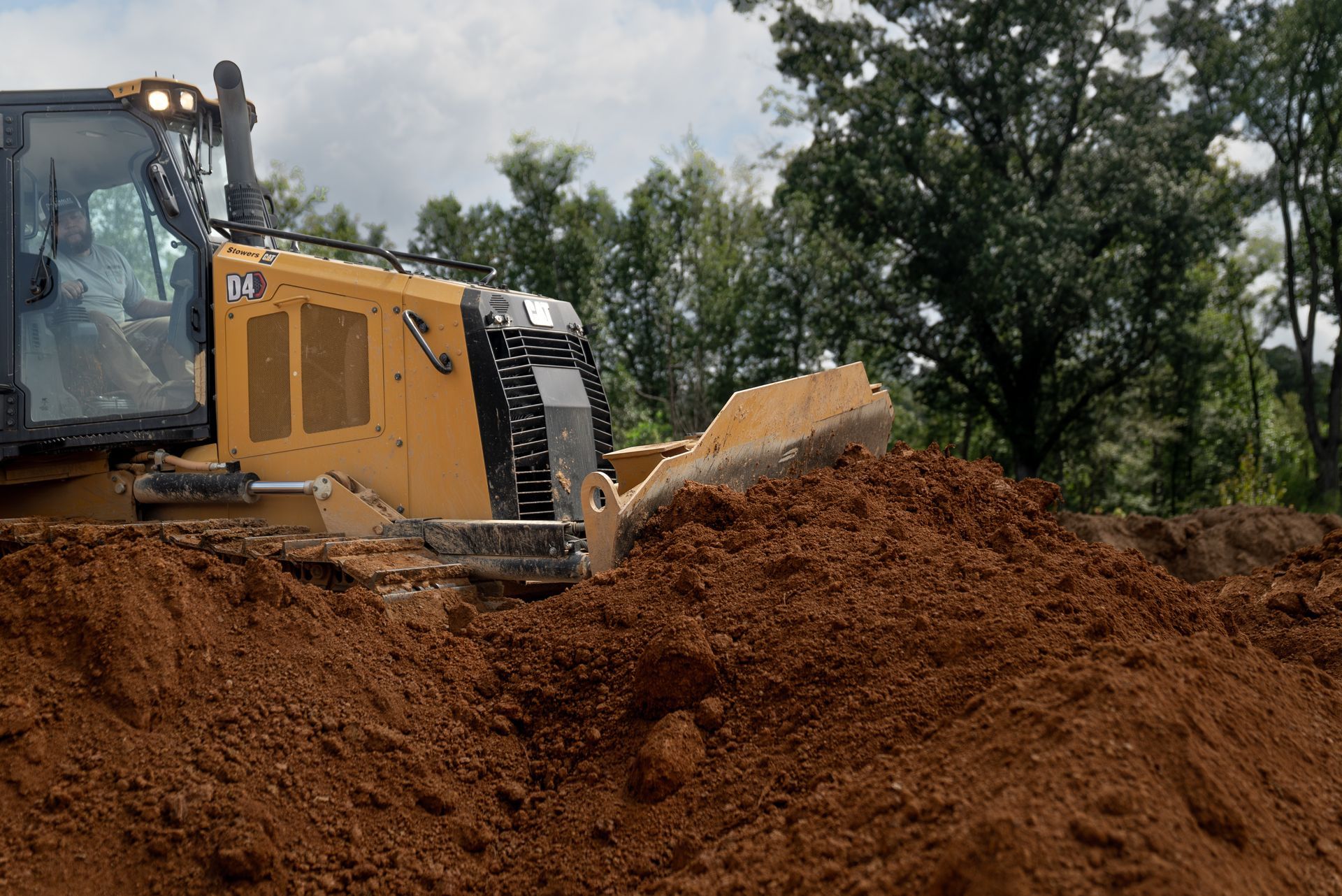 Yellow bulldozer pushing dirt. Operator in cab, trees in background.