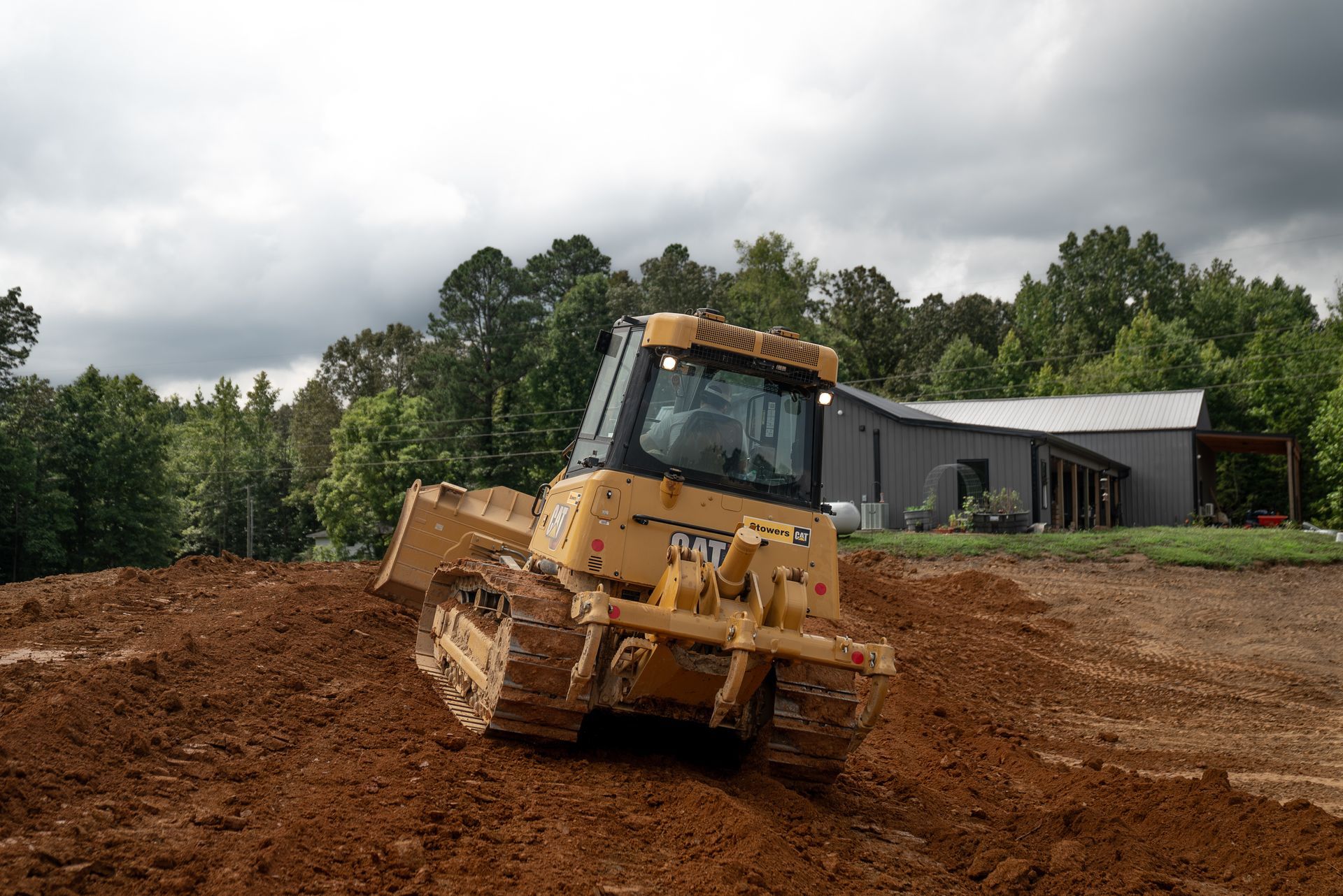 Yellow Caterpillar bulldozer grading a dirt lot near a house under a cloudy sky.
