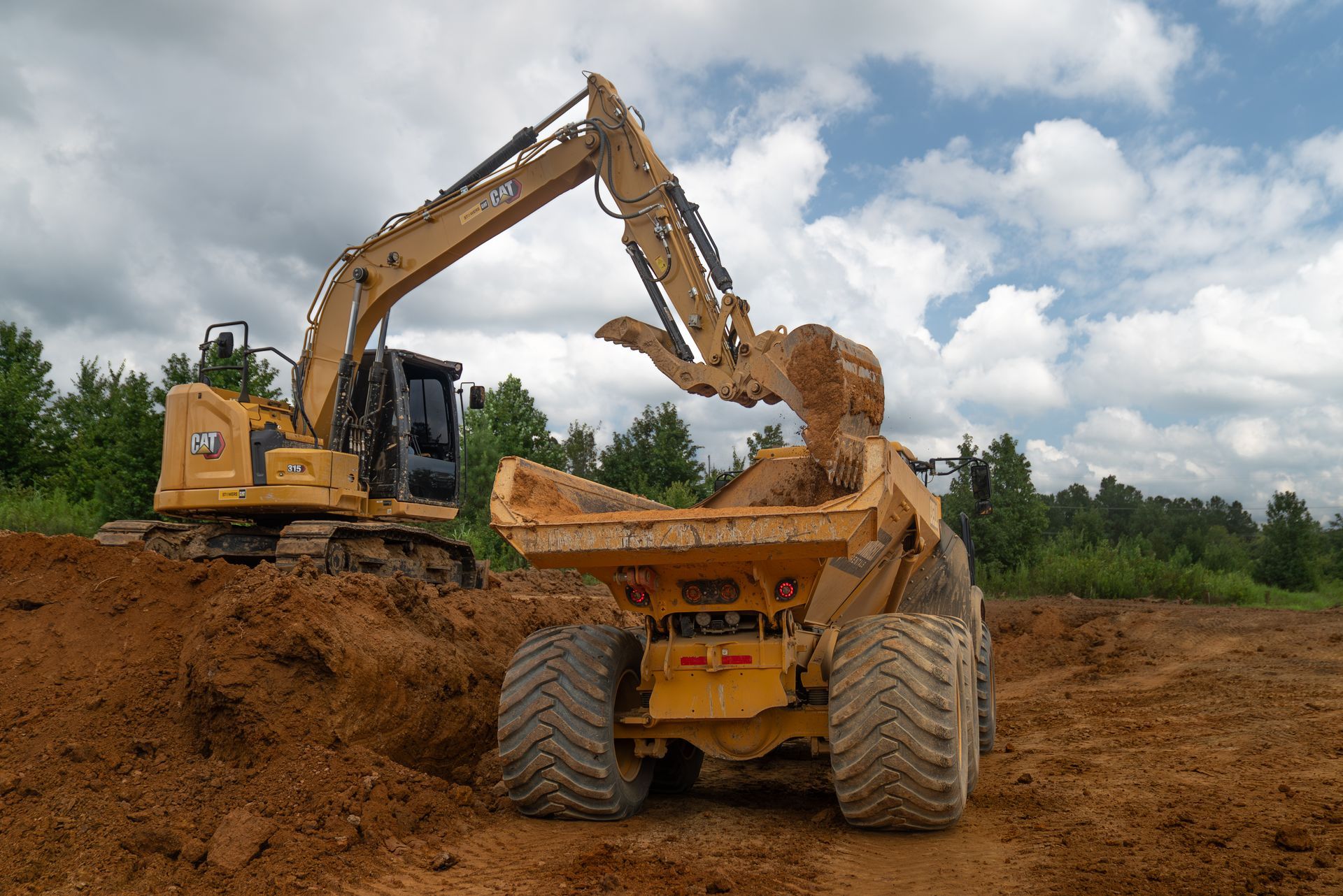 Yellow excavator loading dirt into a yellow dump truck on a construction site under a cloudy sky.