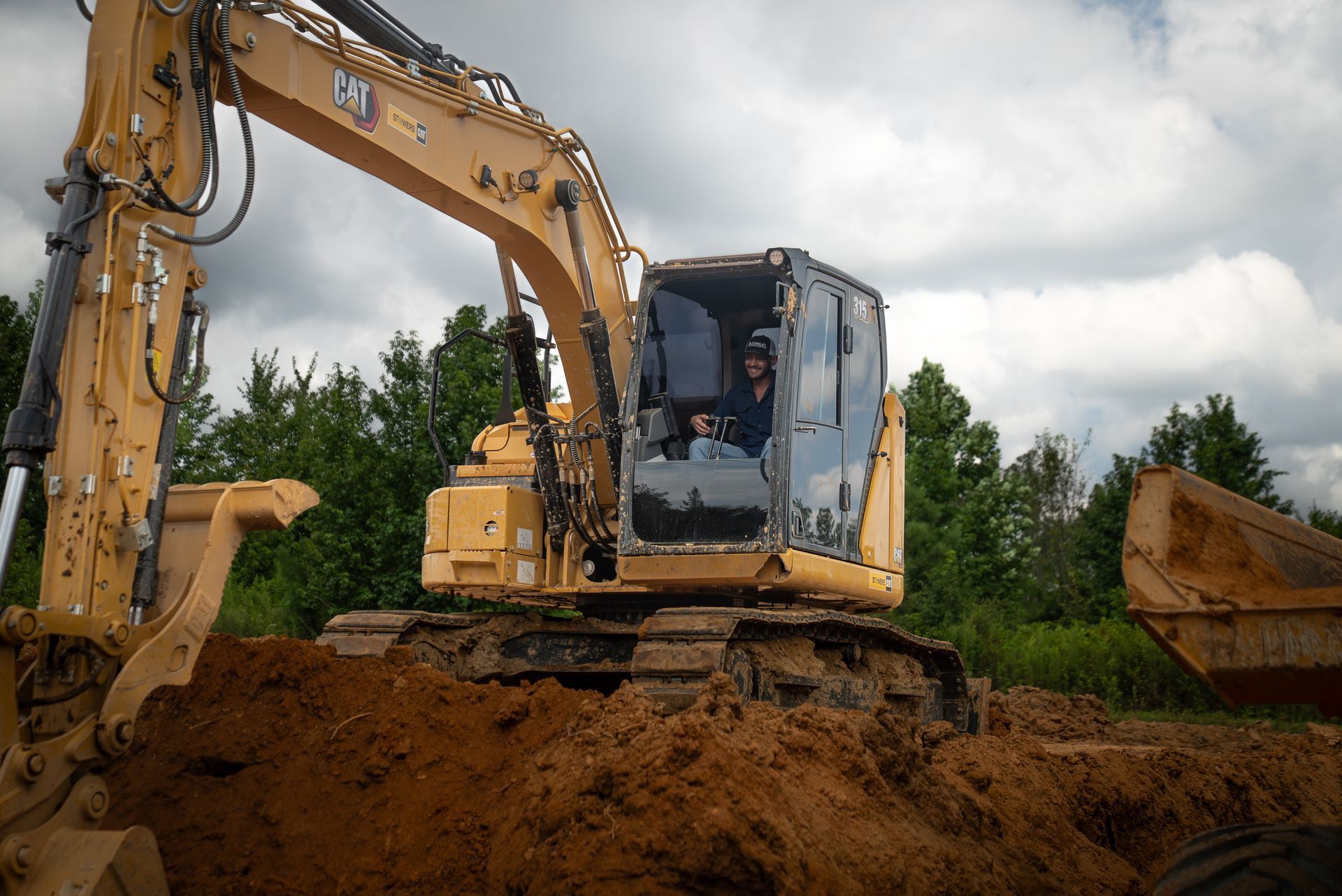 Yellow excavator on a dirt pile, operator visible in the cab. Cloudy sky, trees in the background.