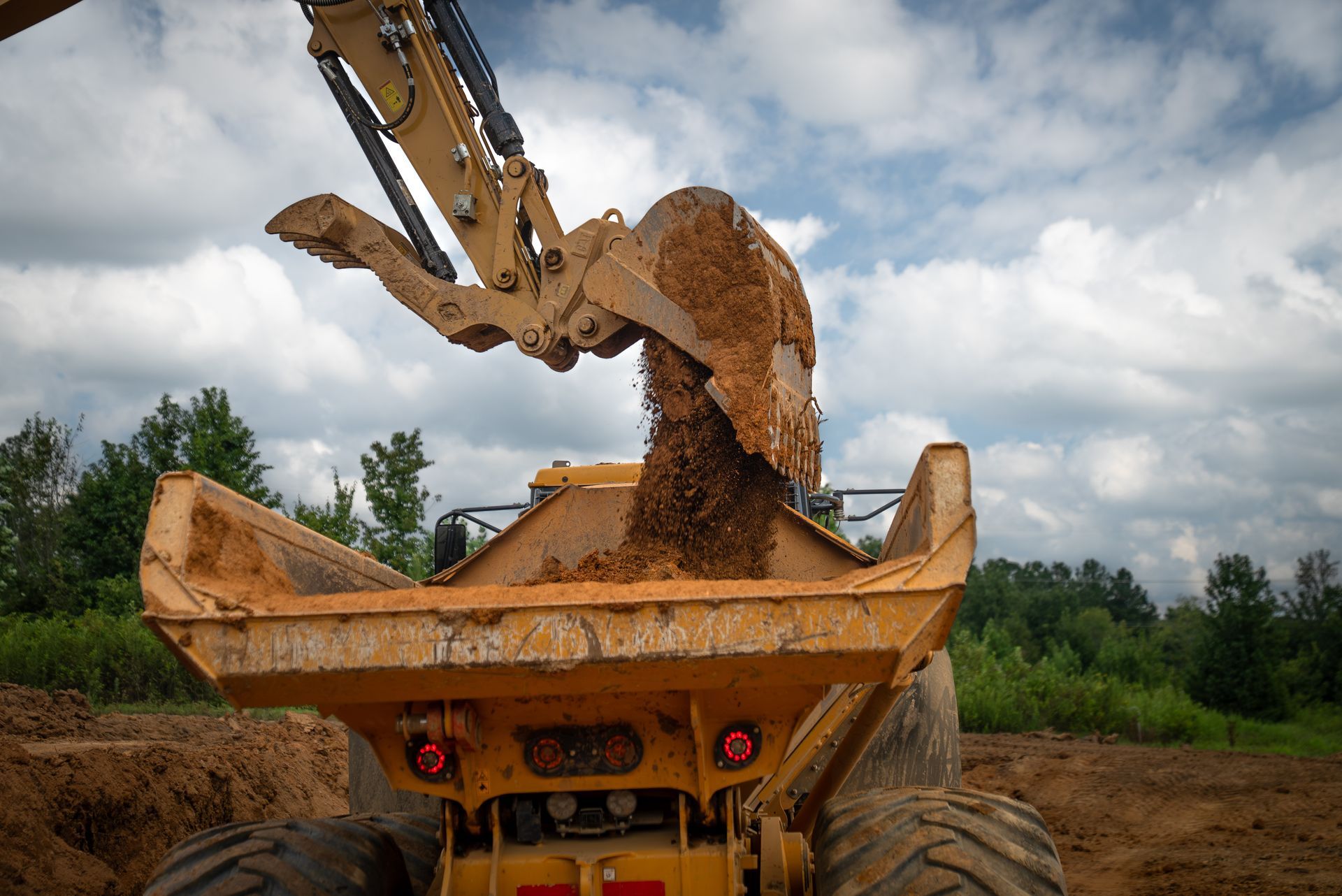 Yellow excavator dumping soil into a yellow dump truck bed at a construction site.