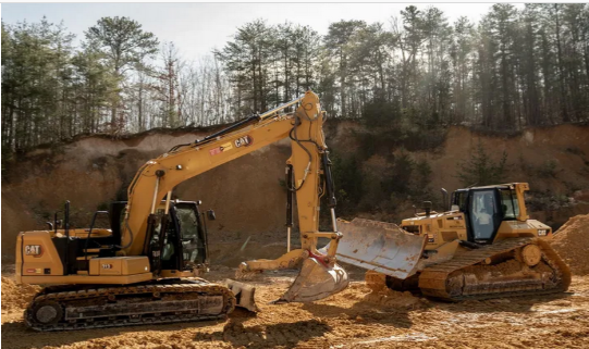 Construction site with dirt, gravel, and trees under a blue sky, an excavator works in the distance.