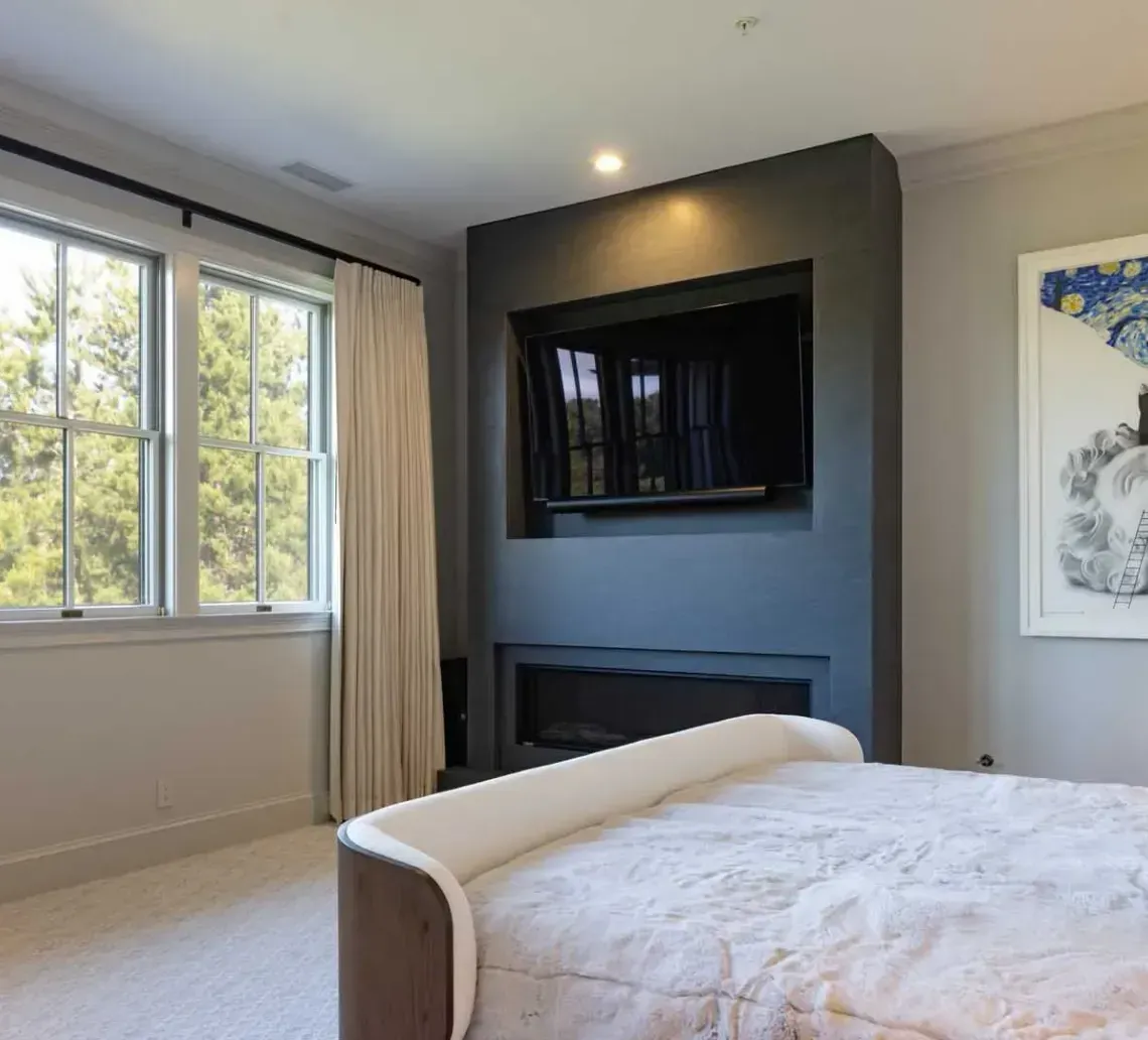 A bedroom with a bed, a dark grey fireplace wall with an integrated television, and framed art on the right.