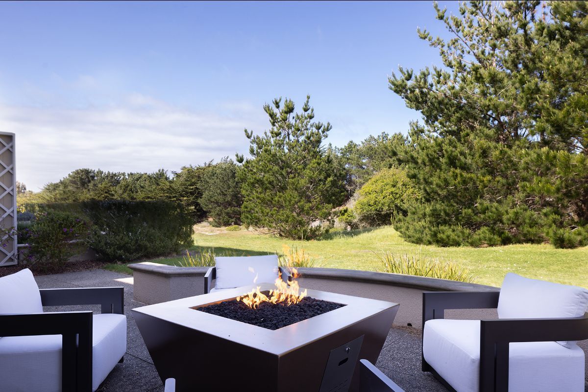 Outdoor fire pit on a patio with white lounge chairs overlooking a grassy lawn and trees on a sunny day.