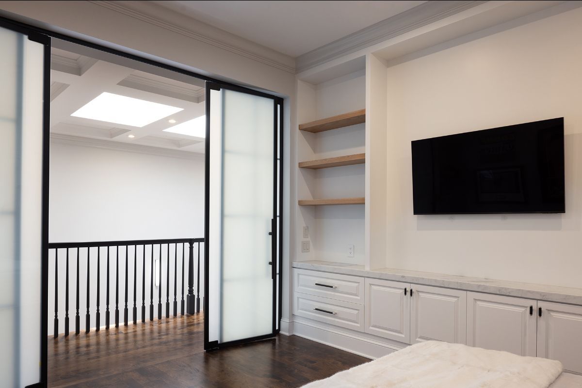A room with white built-in shelving and cabinets, a wall-mounted TV, and frosted glass sliding doors opening to a hallway.