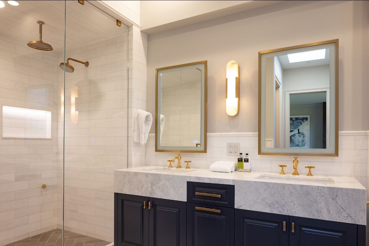 A modern bathroom featuring a double vanity with navy cabinets, a white marble countertop, gold fixtures, and a shower.