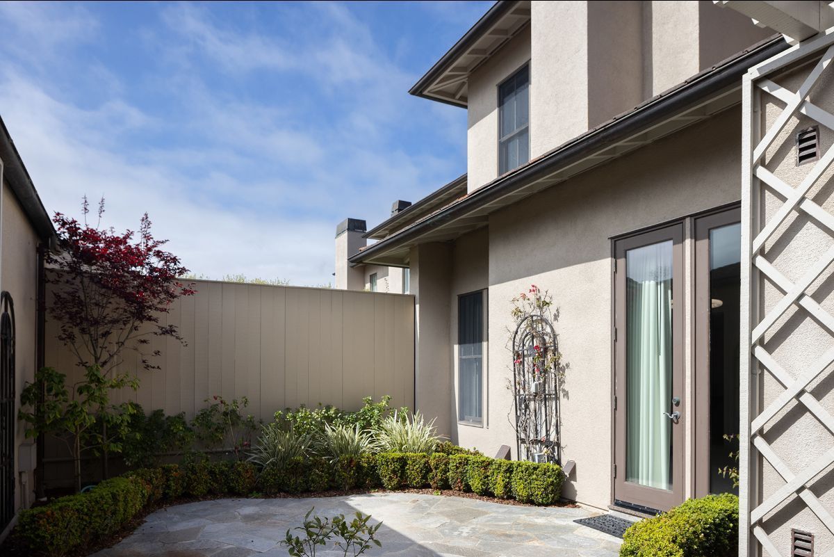 A secluded stone patio area next to a stucco house, featuring a manicured hedge, a decorative trellis, and a glass door.