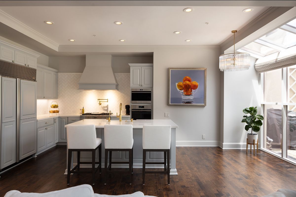 A modern kitchen with grey cabinets, a central island with three white stools, dark wood floors, and a floral painting.