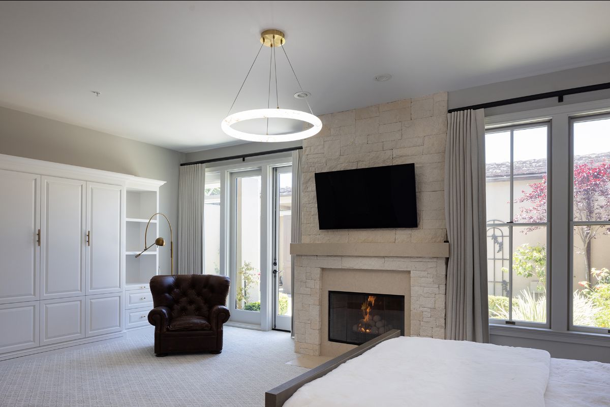 A bright bedroom featuring a stone fireplace with a mounted television, a brown leather armchair, and a circular pendant.