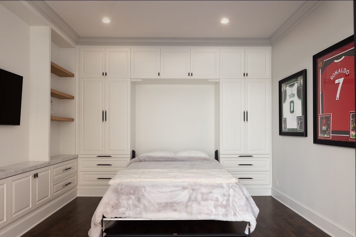A Murphy bed is folded down in a white bedroom with floor-to-ceiling cabinets, open wood shelving, and framed jerseys.