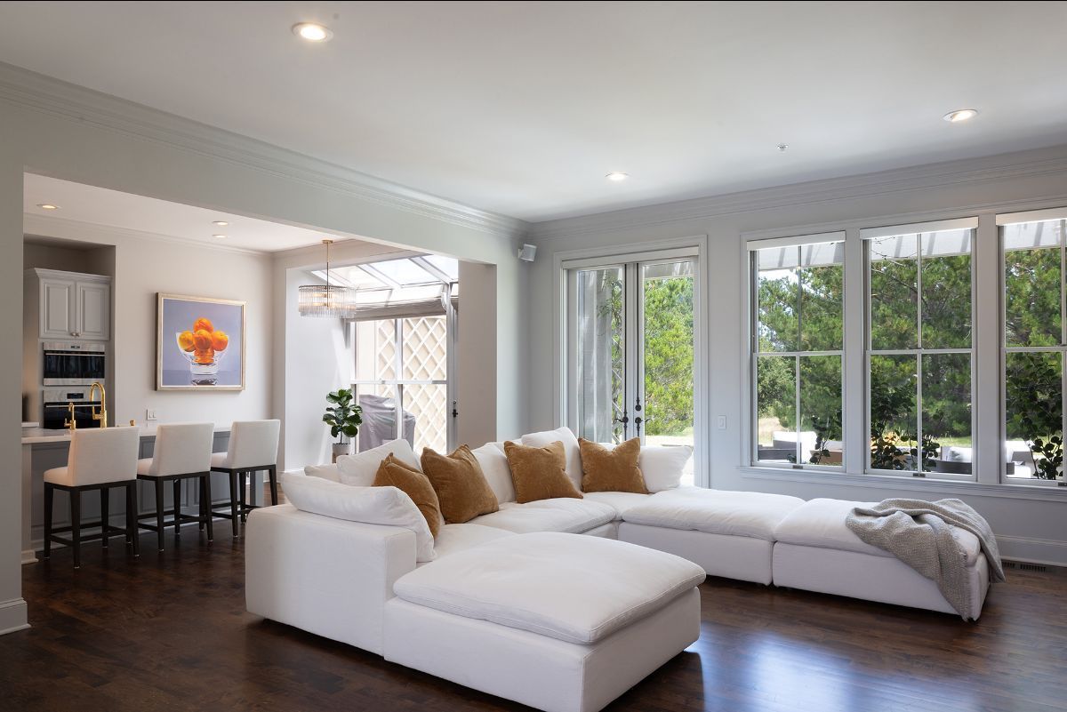 A bright living room with a large white sectional sofa, three bar stools at a counter, and tall windows facing greenery.