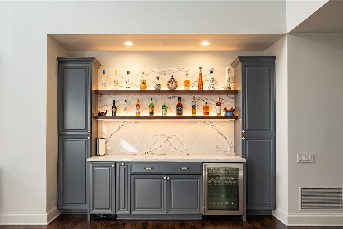 A home bar with charcoal cabinets, marble backsplash, floating shelves holding liquor bottles, and a wine refrigerator.