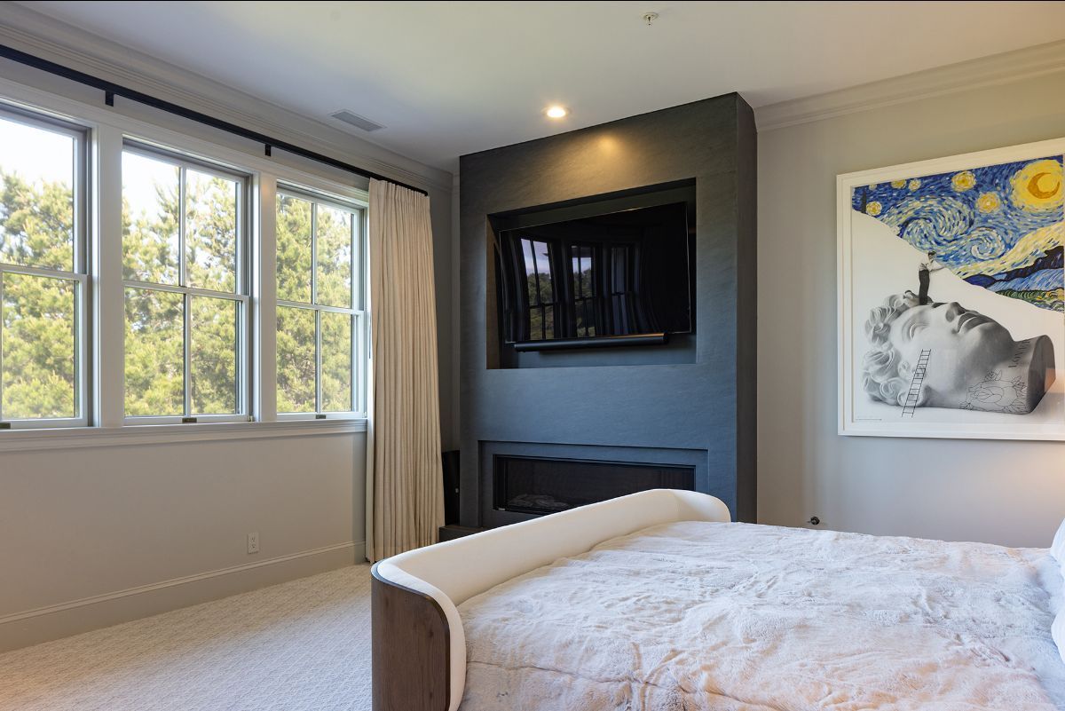 A bedroom featuring a bed, a dark accent wall with a recessed TV and fireplace, large windows with drapes, and wall art.