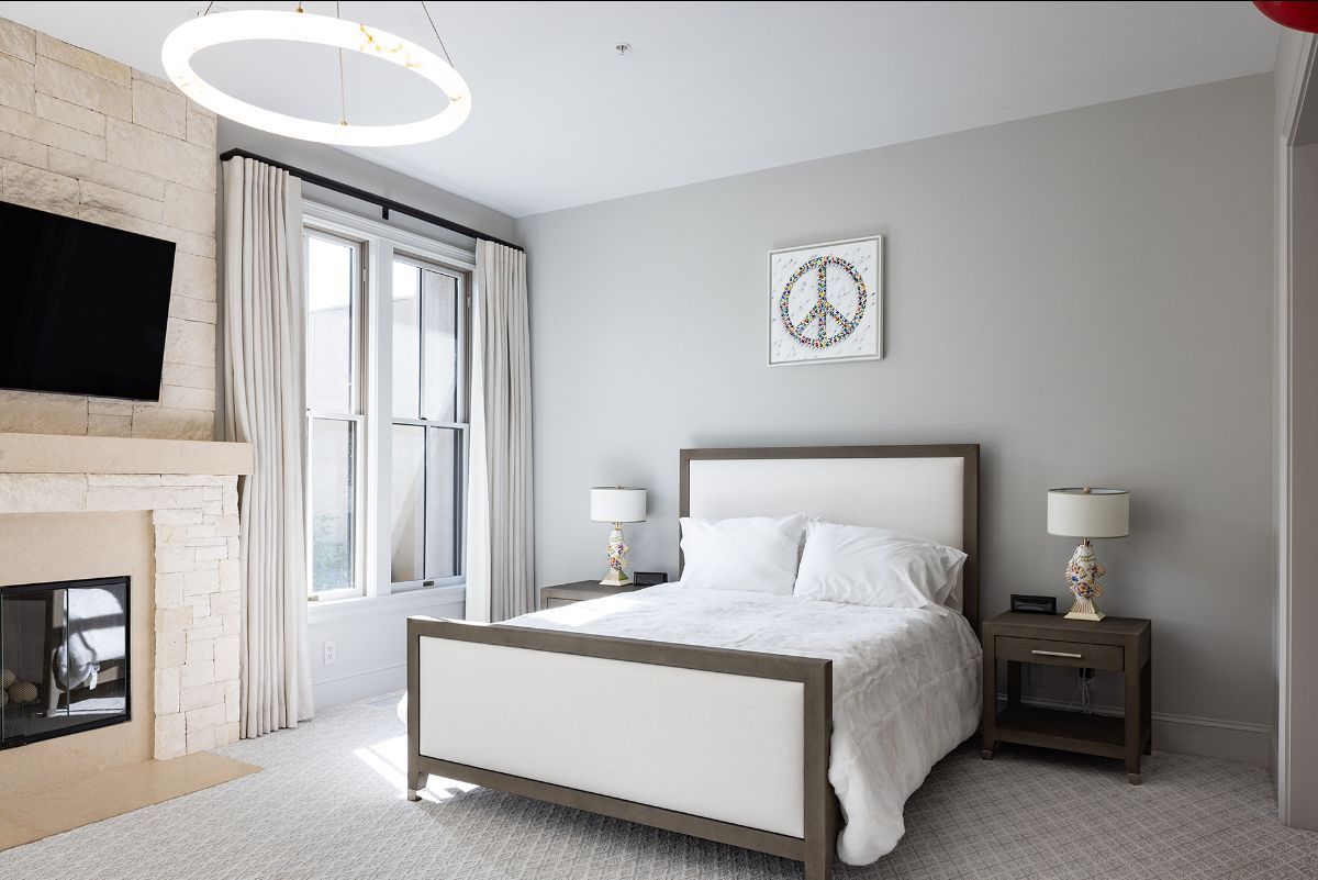 A bright bedroom with a fireplace, a queen bed, two nightstands, lamps, and curtains flanking a window.