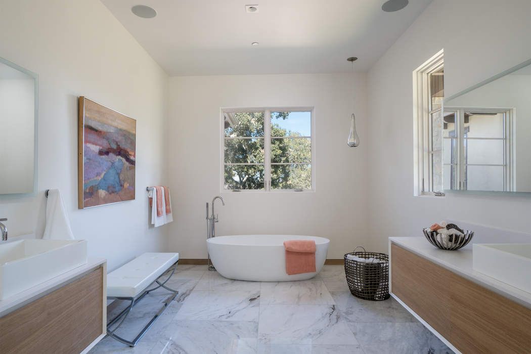 A bright, modern bathroom featuring a white soaking tub, light wood vanities, marble flooring, and a large window.