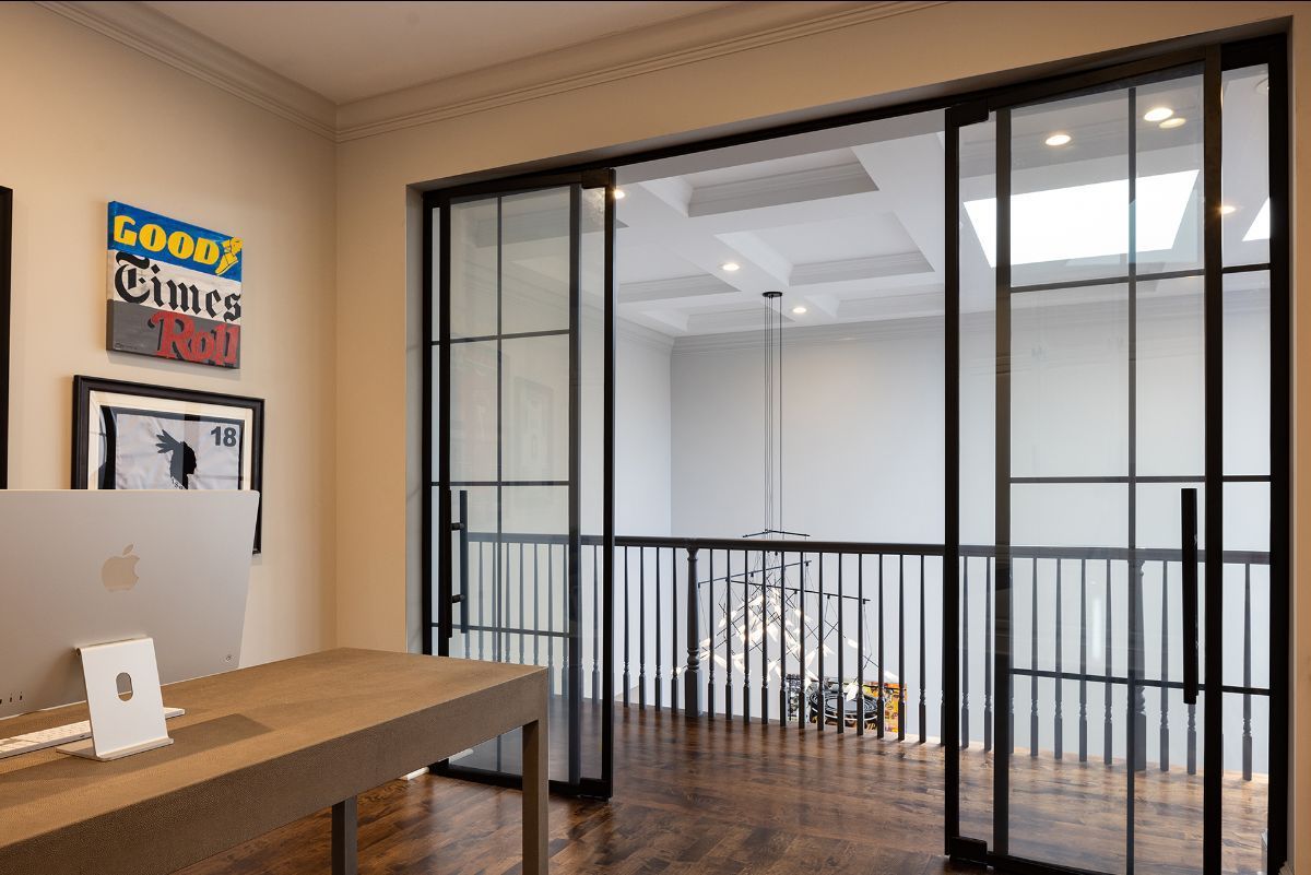 A desk in a home office faces large, black-framed glass sliding doors that look out onto a modern, multi-level hallway.
