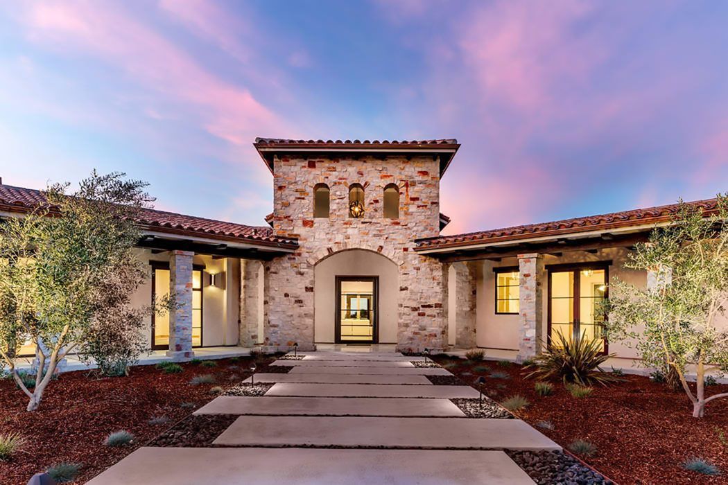 A stone-faced Mediterranean home with a tile roof, centered front door, and walkway set against a vibrant sunset sky.