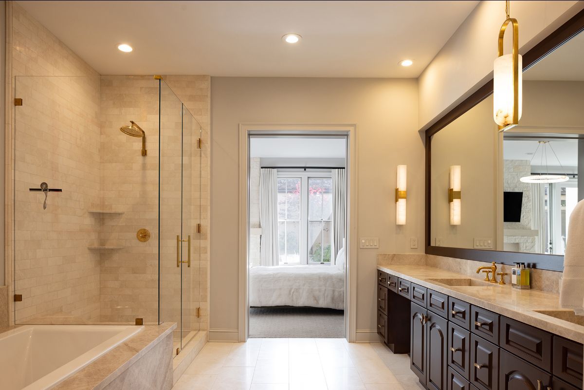 A modern bathroom features a glass-enclosed shower, a dark wood vanity with a large mirror, and a doorway to a bedroom.