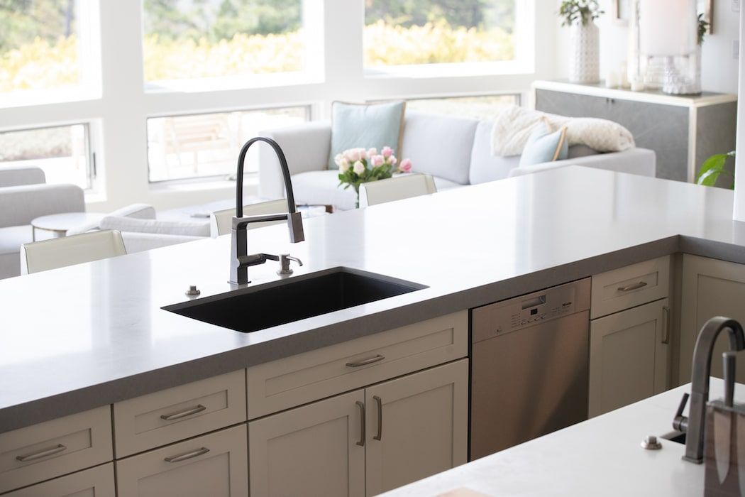 A modern kitchen island with a dark sink, faucet, and dishwasher, overlooking a bright, open-plan living room.