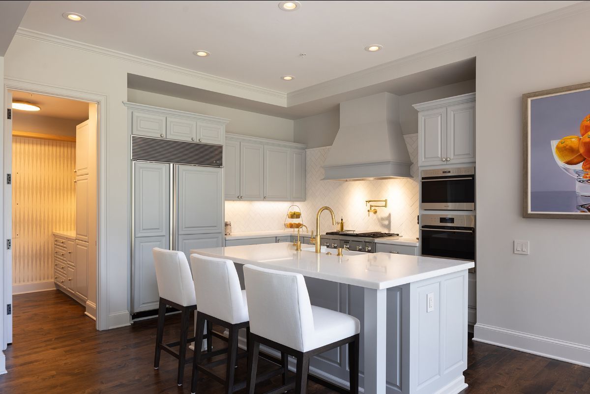 A bright kitchen featuring a white marble island with three stools, grey cabinetry, stainless appliances, and wood floors.