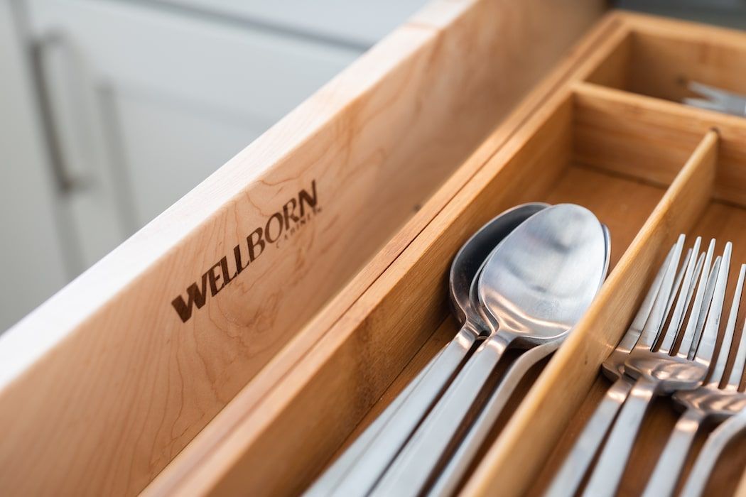 A wooden kitchen drawer with a Wellborn brand logo containing a bamboo silverware organizer filled with metal utensils.