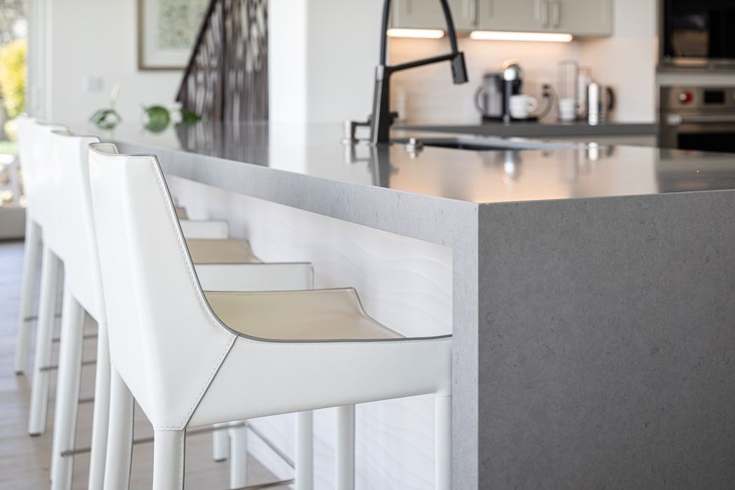 Modern kitchen island with gray stone countertop and white leather bar stools.