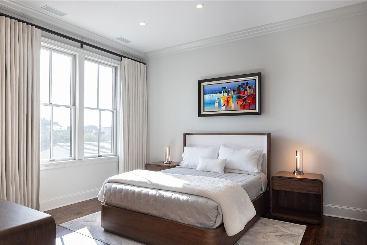 A modern, sunlit bedroom featuring a wooden bed frame, white bedding, two bedside tables, and a vibrant framed painting.