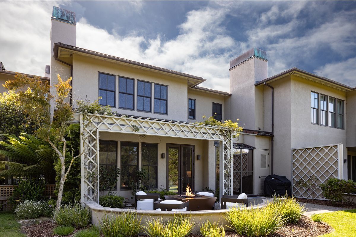 A two-story beige house with a latticed patio pergola, outdoor seating, and a fire pit in a sunny garden setting.