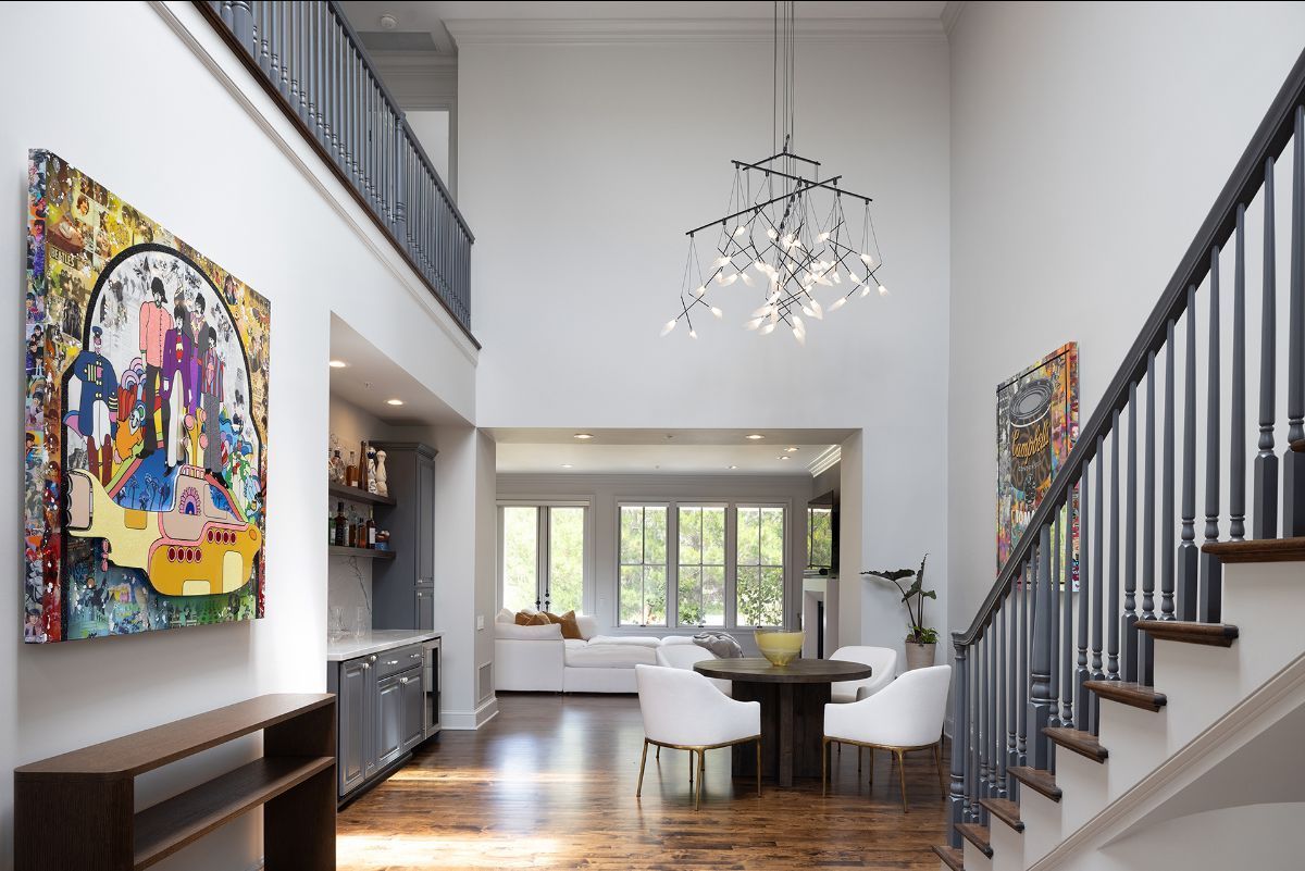 A high-ceilinged modern home entryway with a central chandelier, staircase, dining area, and large colorful wall art.
