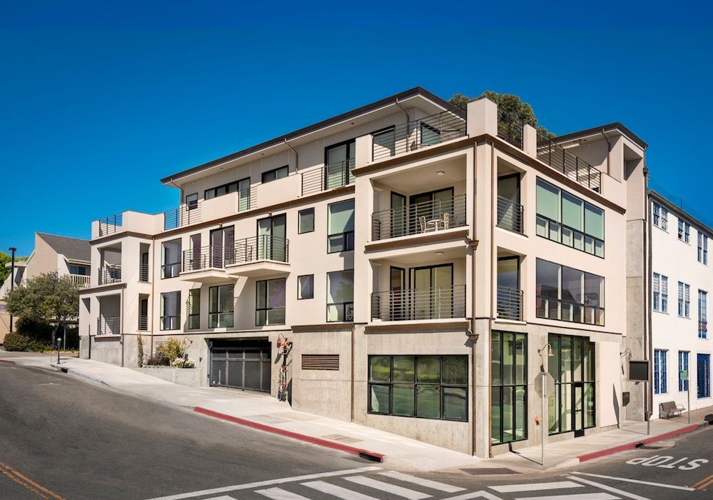 A multi-story beige apartment building with balconies, glass windows, and a ground-floor garage on a sunny street corner.