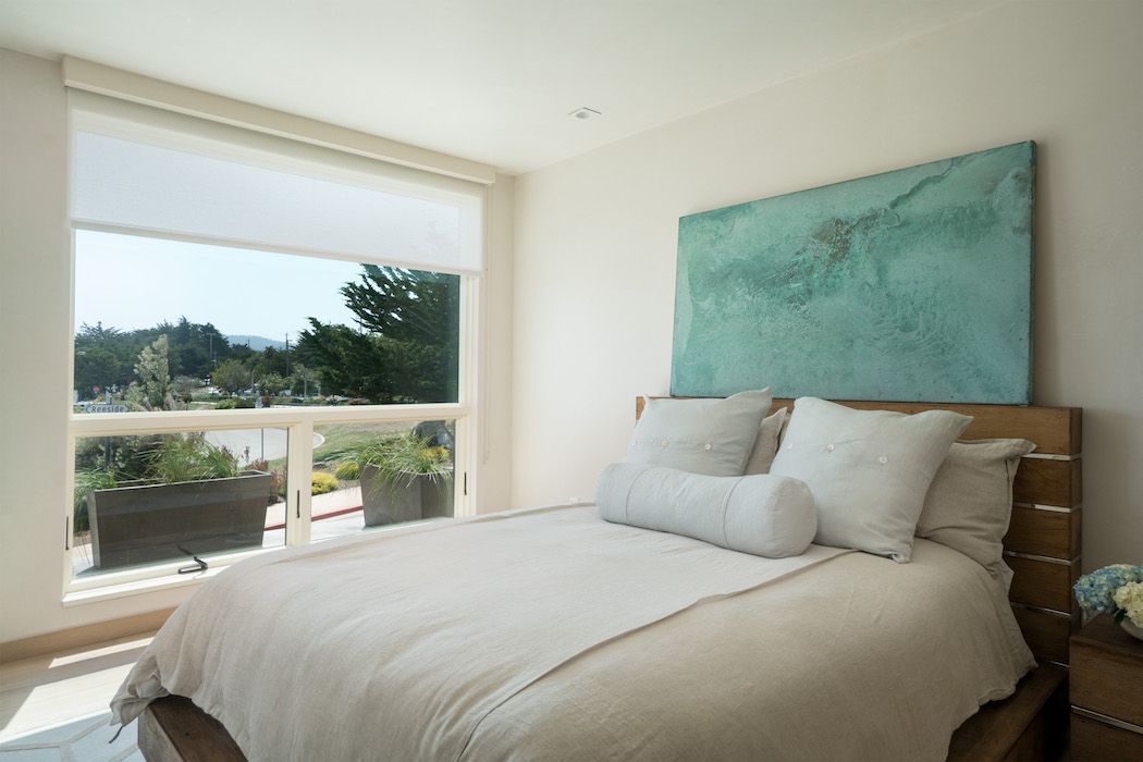 Modern bedroom featuring a bed with white bedding, a turquoise abstract art piece, and a large window with garden views.