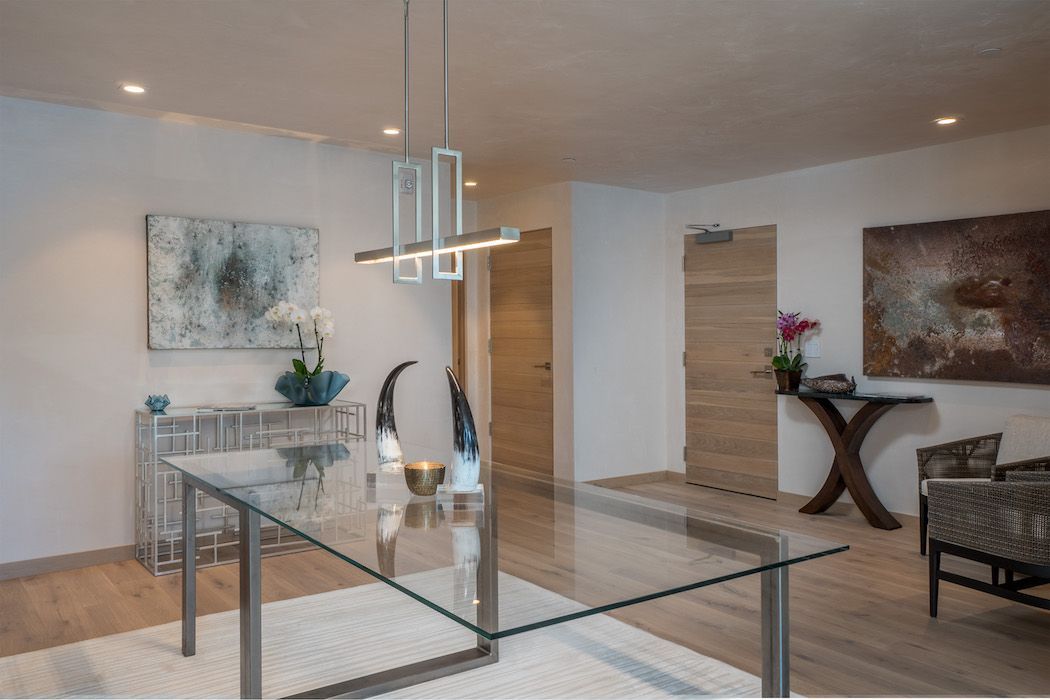 A modern interior space with a glass table, contemporary light fixture, two wooden doors, and abstract wall art.